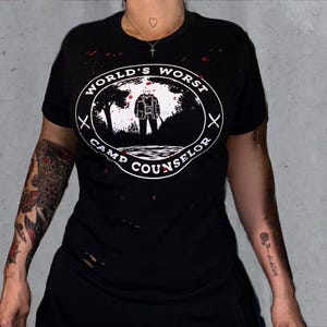 May include: Black t-shirt with the text "WORLD'S WORST CAMP COUNSELOR" in a white oval design. The design features a figure in a mask standing in front of a forest scene. The shirt has distressed details and red splatter.