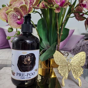 May include: A dark brown bottle of "PRE-POO Café / Coffe" with a black pump dispenser, a gold butterfly decoration, and a bouquet of orchids with purple and yellow spotted petals. The bottle has a white label with a woman's silhouette.
