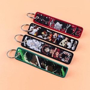 Könnte beinhalten: Vier rechteckige, bestickte Schlüsselanhänger mit Metallringen. Jeder Schlüsselanhänger hat ein anderes farbenfrohes Design im Anime-Stil. Die Schlüsselanhänger haben rote, schwarze und grüne Ränder. Der Hintergrund ist hellorange.