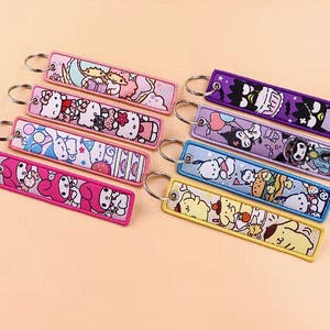 Puede incluir: Colección de llaveros bordados coloridos con personajes de dibujos animados. Cada llavero rectangular tiene un anillo de metal y un diseño único, incluyendo Hello Kitty y otros personajes, en tonos de rosa, morado, azul y amarillo.