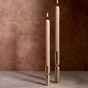 May include: Two lit, slender, blush-colored candles in brass holders. The candles have a classic taper shape and are set against a textured, reddish-brown background. The holders have a unique hourglass design.