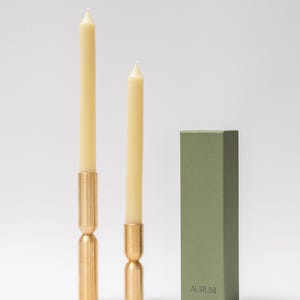 May include: Two cream-colored taper candles with brass holders and a green box. The candles are of different heights, with the taller one on the left. The brass holders have a unique hourglass shape. The green box has the word "AURUM" printed on it.