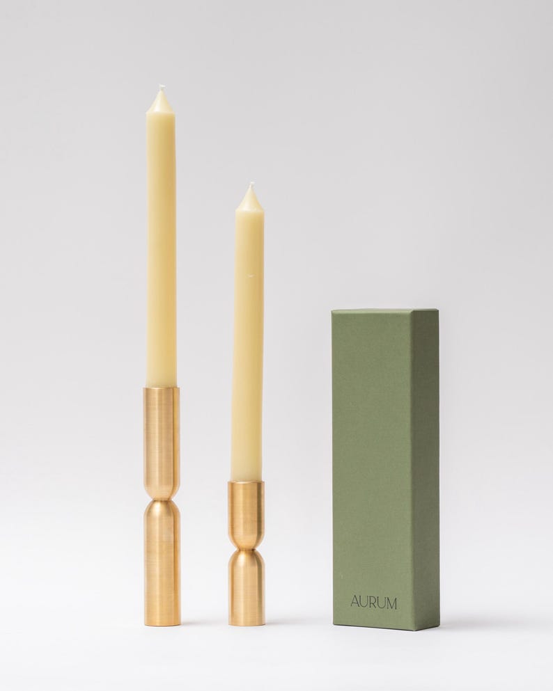 May include: Two cream-colored taper candles with brass holders and a green box. The candles are of different heights, with the taller one on the left. The brass holders have a unique hourglass shape. The green box has the word "AURUM" printed on it.