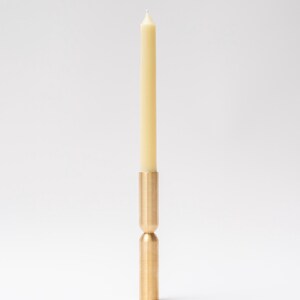 May include: A brass candle holder with a cream-colored taper candle. The candle holder has a cylindrical base and a matching top, connected by a rounded midsection. The candle is centered and stands upright.