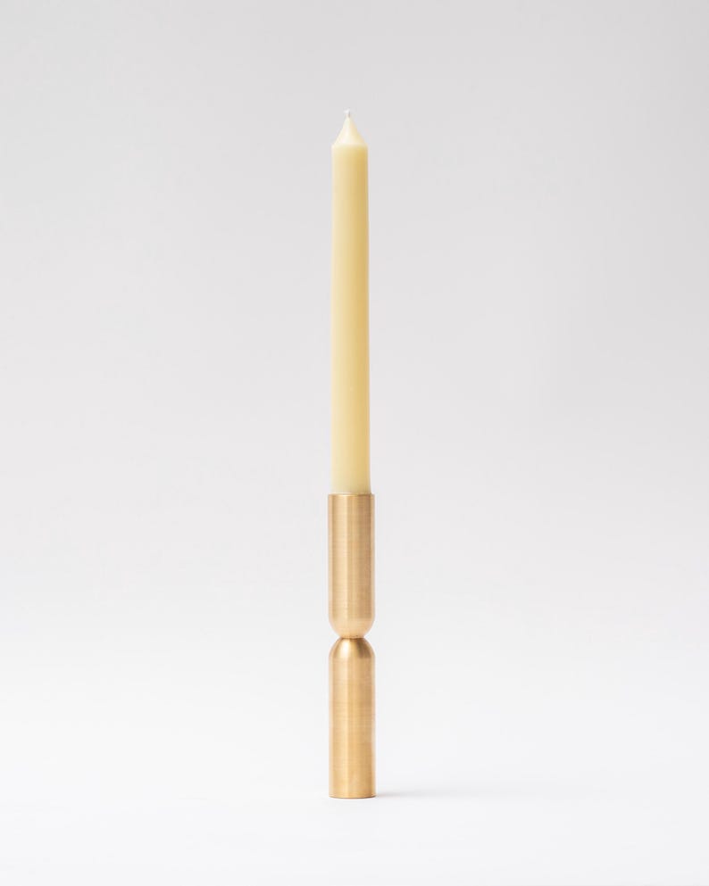 May include: A brass candle holder with a cream-colored taper candle. The candle holder has a cylindrical base and a matching top, connected by a rounded midsection. The candle is centered and stands upright.