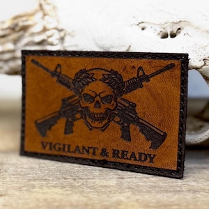 May include: A rectangular brown leather patch with a skull and crossed rifles design. The skull is centered, with a laurel wreath above it. The text "VIGILANT & READY" is below the skull. The patch has a black border.