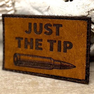 May include: A rectangular brown leather patch with the words "JUST THE TIP" embossed in dark brown letters. Below the text is an illustration of a bullet. The patch has a dark brown border.