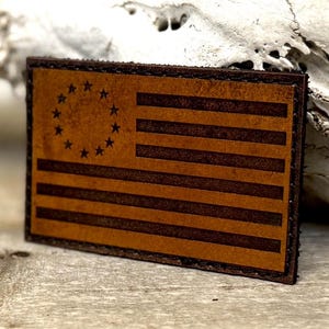 May include: A rectangular leather patch featuring a brown and tan design of the American flag. The flag has 13 stripes and a circle of 13 stars. The patch has a stitched border.