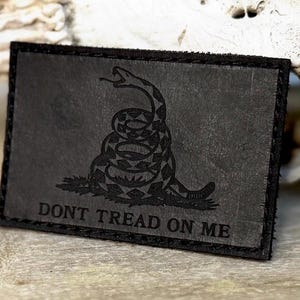May include: A rectangular black patch with a stitched border. The patch features a coiled snake illustration and the words "DONT TREAD ON ME" in capital letters. The snake is in a striking, detailed design.