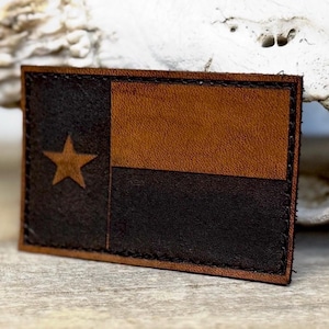 May include: A rectangular leather patch featuring the Texas state flag design. The patch is divided into three sections: a dark brown section with a gold star, a light brown section, and a black section. The edges are stitched with black thread.