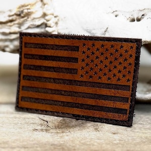 May include: A rectangular, brown leather patch featuring the American flag design. The flag has dark brown stripes and a field of stars. The patch is likely for a jacket or bag.