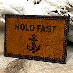 May include: A rectangular, brown leather patch with the words "HOLD FAST" embossed across the top. An anchor is embossed below the text. The patch has a dark brown border with decorative stitching.