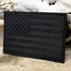 May include: A rectangular, black leather patch featuring a debossed American flag design. The flag's stars and stripes are subtly raised, and the patch has a stitched border. The overall aesthetic is a subdued, monochromatic look.