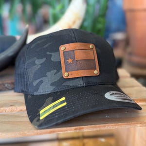 May include: Black camouflage trucker hat with a leather patch featuring the Texas flag. The hat has a curved brim and a mesh back. The patch is secured with brass rivets. A yellow sticker with the word "MULTICAM" is on the brim.