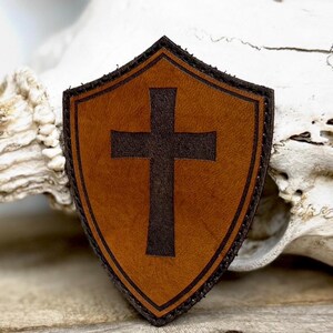 May include: A shield-shaped leather patch with a dark brown cross on a tan background. The patch has a dark brown border and stitching. The patch is likely a religious symbol.