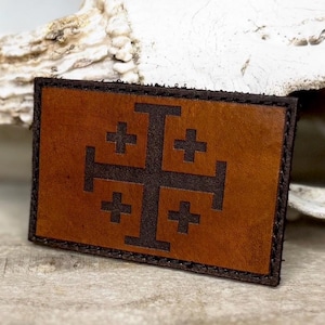 May include: A rectangular brown leather patch with a dark brown Jerusalem cross design. The patch has a stitched border and is set against a blurred background.