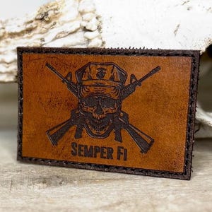 May include: A rectangular brown leather patch with a skull wearing a cap and crossed rifles. The text "Semper Fi" is below the skull. The patch has a dark brown border and is likely a military-themed accessory.
