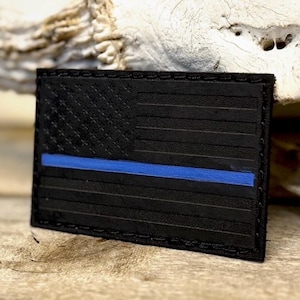 May include: A black rectangular patch featuring the American flag design with a blue stripe. The flag's stars and stripes are subtly embossed on the patch's surface. The patch is likely made of leather or a similar material.