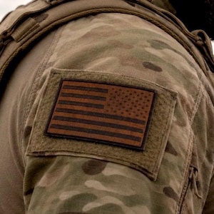 May include: A rectangular patch featuring a brown and black United States flag design attached to a camouflage patterned sleeve. The flag patch is centered on a matching camouflage fabric panel with visible stitching along the edges of the patch.
