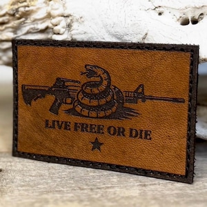 May include: A rectangular brown leather patch with a black border. The patch features a detailed image of a coiled snake over a rifle and the text "LIVE FREE OR DIE" with a star below.
