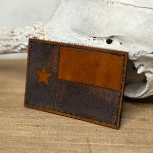 May include: A rectangular leather patch featuring the Texas flag design. The patch has a brown and dark brown color scheme with a star in the upper left corner. The edges are stitched with black thread.