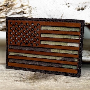 May include: A rectangular patch featuring the American flag design. The flag has a brown leather border and a wooden appearance. The stripes alternate between camouflage and brown, with stars in the upper left corner.