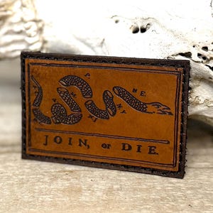 May include: A rectangular, brown leather patch with a dark brown border. The patch features a segmented snake illustration and the words "JOIN, or DIE" in dark brown lettering. The patch is likely a decorative item.