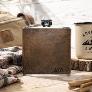 May include: A brown leather-wrapped flask with a silver cap and trim. The initials "AKO" are engraved on the front. A canvas bag, plaid fabric, and a camping mug with the words "Adventure Awaits" are in the background.