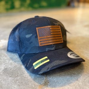May include: A navy blue camouflage baseball cap with a brown leather American flag patch. The cap has a mesh back and a curved brim. A yellow and black sticker is on the brim.
