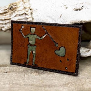 May include: A rectangular leather patch with a brown background and a black border. The patch features a green and brown camouflage design of a pirate flag with a skeleton holding a weapon and a heart pierced by an arrow. The patch is likely for a jacket or bag.