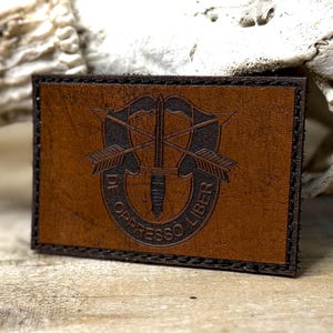 May include: A rectangular brown leather patch featuring a laser-engraved Special Forces insignia. The design includes a dagger centered between two crossed arrows, enclosed by a curved banner with the text DE OPPRESSO LIBER. The edges are stitched.