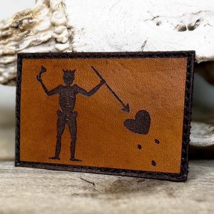 May include: A rectangular brown leather patch with a black border. The patch features a black silhouette of a figure holding a spear and a goblet, with a heart pierced by an arrow. The design is reminiscent of a pirate flag.