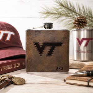 May include: A brown leather flask with the letters "VT" in black. The flask is next to a maroon baseball cap and a silver mug with the same "VT" logo. A small notebook, pen, and keychain are also visible.
