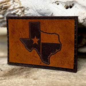May include: A rectangular leather patch featuring the outline of the state of Texas. The Texas shape is divided into two sections, one brown and one dark brown with a star. The patch has a brown border and is likely a decorative item.