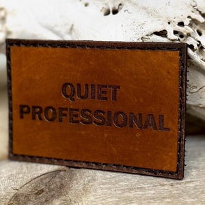 May include: A rectangular, brown leather patch with the words "QUIET PROFESSIONAL" embossed in dark brown. The patch has a stitched border and a slightly distressed appearance, suggesting a vintage or handcrafted style.