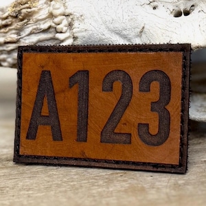 May include: A rectangular leather patch with the letters "A123" embossed in dark brown. The patch has a light brown center and a dark brown stitched border. The background is a light brown wooden surface.