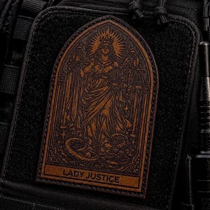 May include: A brown leather patch featuring Lady Justice, holding scales and a sword, with a crown and a serpent at her feet. The patch is rectangular with an arched top and the words "LADY JUSTICE" at the bottom. The patch is attached to a black fabric.