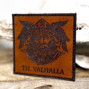 May include: Square, brown leather patch with a detailed image of a Viking warrior wearing a helmet and surrounded by ravens. The text "TIL VALHALLA" is embossed at the bottom. The patch has a dark brown border.