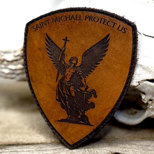 May include: Shield-shaped leather patch with the words "SAINT MICHAEL PROTECT US" and an image of an angel holding a cross and sword. The patch is brown with a dark brown border and image.