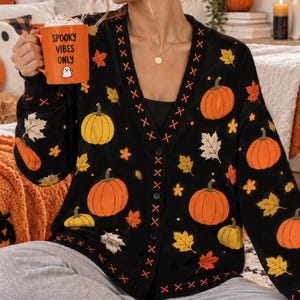 May include: A black V-neck cardigan featuring a repeating pattern of orange and yellow pumpkins and autumn leaves, with orange cross-stitch detailing along the front placket and hem, and a row of black buttons down the center front.