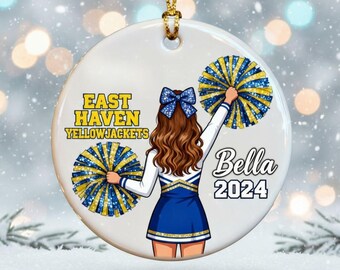 May include: A white ceramic ornament with a gold string for hanging. The ornament features a cheerleader with brown hair, a blue bow, and a blue and white uniform. Text includes "East Haven Yellowjackets" and "Bella 2024".