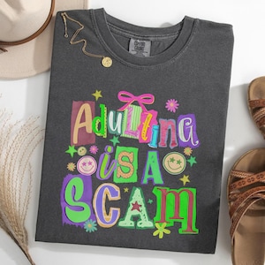 以下が含まれることがあります： 「Adulting is a Scam」というフレーズがカラフルで気まぐれな文字で書かれた、濃いグレーのComfort Colors Tシャツ。デザインには星、花、スマイリーフェイスが含まれています。ゴールドのネックレスとサンダルが見えます。