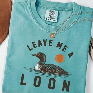 May include: A teal short-sleeved t-shirt featuring a graphic of a loon swimming on water with an orange sun. The text "LEAVE ME A LOON" is printed in dark brown capital letters. The shirt has a Comfort Colors brand label at the inner neckline.