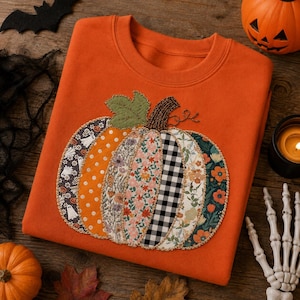 May include: An orange crewneck sweatshirt featuring a large, central pumpkin applique. The pumpkin is constructed from various fabric patches, including ghost prints, orange polka dots, floral patterns, and black and white gingham checks.