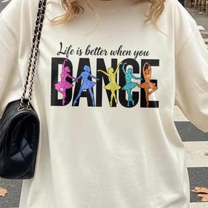 May include: A cream-coloured t-shirt with the text "Life is better when you DANCE." The word "DANCE" features colourful glittery ballet dancers in pink, blue, yellow, teal, and orange. The shirt is a casual, comfortable style.