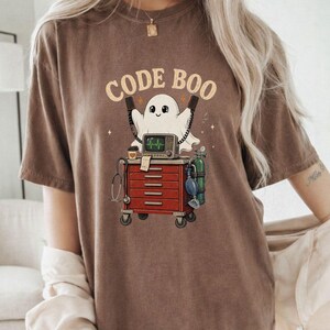 Puede incluir: Una camiseta marrón de manga corta con un gráfico de un fantasma blanco detrás de un carro médico rojo. El carro contiene un monitor, un estetoscopio y un tanque de oxígeno. El texto CODE BOO aparece en una fuente beige arqueada sobre el fantasma.