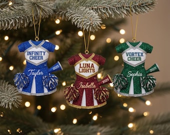 May include: Three cheerleader dress ornaments in blue, maroon, and green, each with glitter accents. The blue ornament says "Infinity Cheer" and "Taylor." The maroon ornament says "Luna Lights" and "Isabella." The green ornament says "Vortex Cheer" and "Sophia."