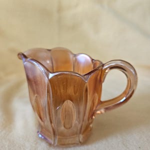 May include: An iridescent amber-colored creamer with a handle and a spout. The creamer has a scalloped rim and a geometric design with oval shapes. The glass has a shiny, reflective surface.