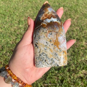 May include: A polished, elongated stone with a pointed top. The stone features a mix of colors, including brown, white, and gray, with speckled and marbled patterns. The stone is held in a hand against a backdrop of green grass.