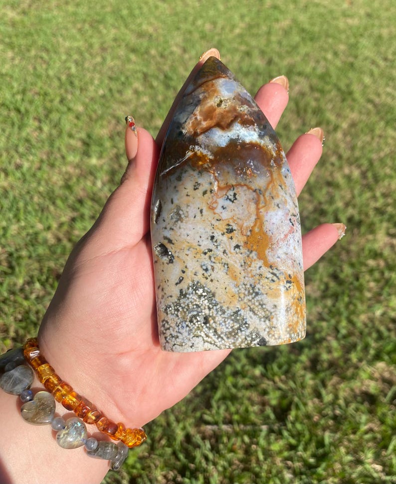 May include: A polished, elongated stone with a pointed top. The stone features a mix of colors, including brown, white, and gray, with speckled and marbled patterns. The stone is held in a hand against a backdrop of green grass.